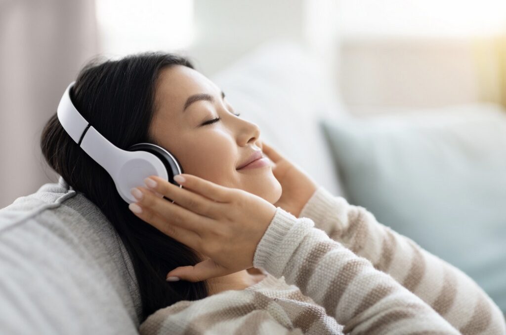 a woman wearing headphones, leaning back on a couch, smiling with her eyes closed as she holds the headphones to her ears