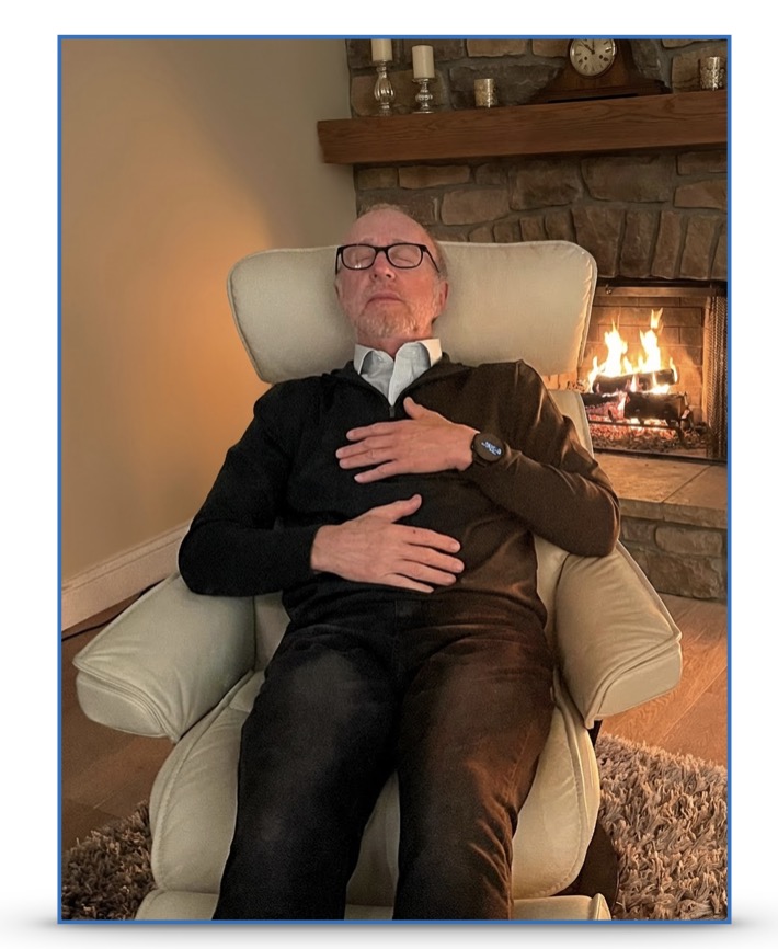 A man sitting back in an armchair, one hand on his heart and one hand on his abdomen